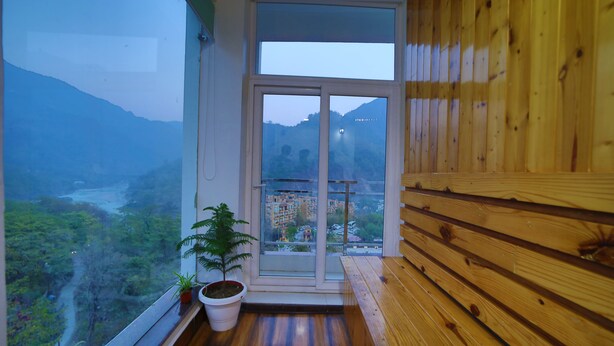 A balcony with wooden flooring, a potted plant, and a view of a mountainous landscape through a glass door. - Kamay The Grand Alova (Narendranagar)