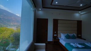 A hotel room with a large window offering a view of a mountainous landscape. The room features a bed with a wooden headboard, a bedside table, and a nightstand. - Kamay The Grand Alova (Narendranagar)