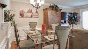 Un coin repas avec une table en verre, des chaises et un lustre. On y trouve un buffet sur lequel est posée une plante, ainsi qu’un tableau accroché au mur.