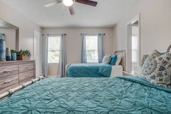 Un dormitorio con cama, cómoda, ventilador de techo y dos ventanas con cortinas.