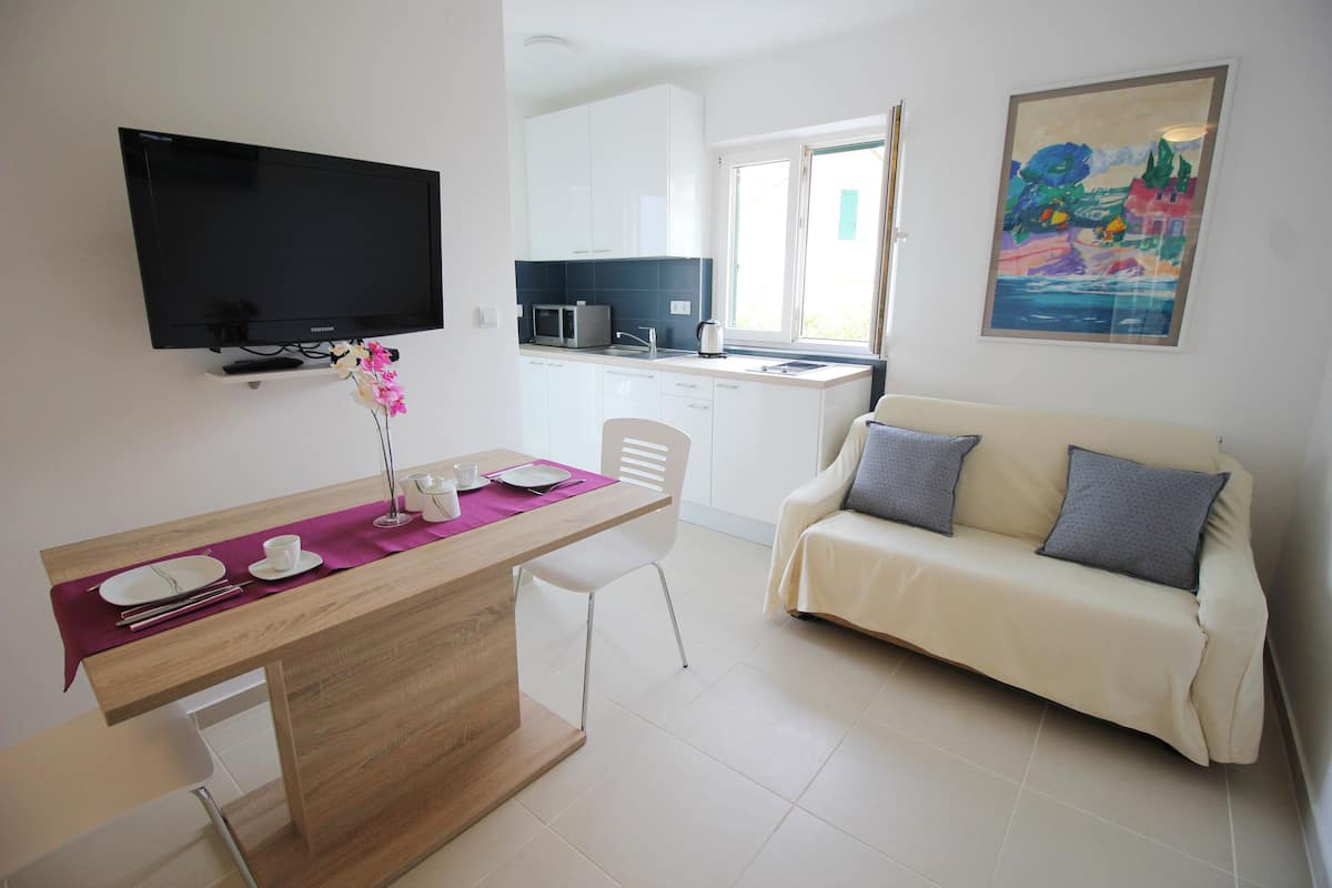 A modern kitchen with white cabinets, a microwave, and a window. There is a dining area with a wooden table, white chairs, and a purple table runner. A beige sofa with gray cushions is against the wall.