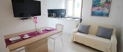 Una cocina moderna con armarios blancos, un microondas y una ventana. Hay una zona de comedor con una mesa de madera, sillas blancas y un mantelí de color púrpura. Un sofá beige con cojines grises está apoyado contra la pared.