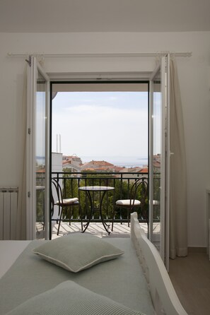A balcony with a table and chairs, a bed with pillows, and a view of the city and sea.