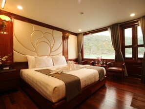 A spacious bedroom with a large bed, wooden flooring, a view of the outdoors, and a nightstand with a lamp. - Calypso Cruises (Tuan Chau)