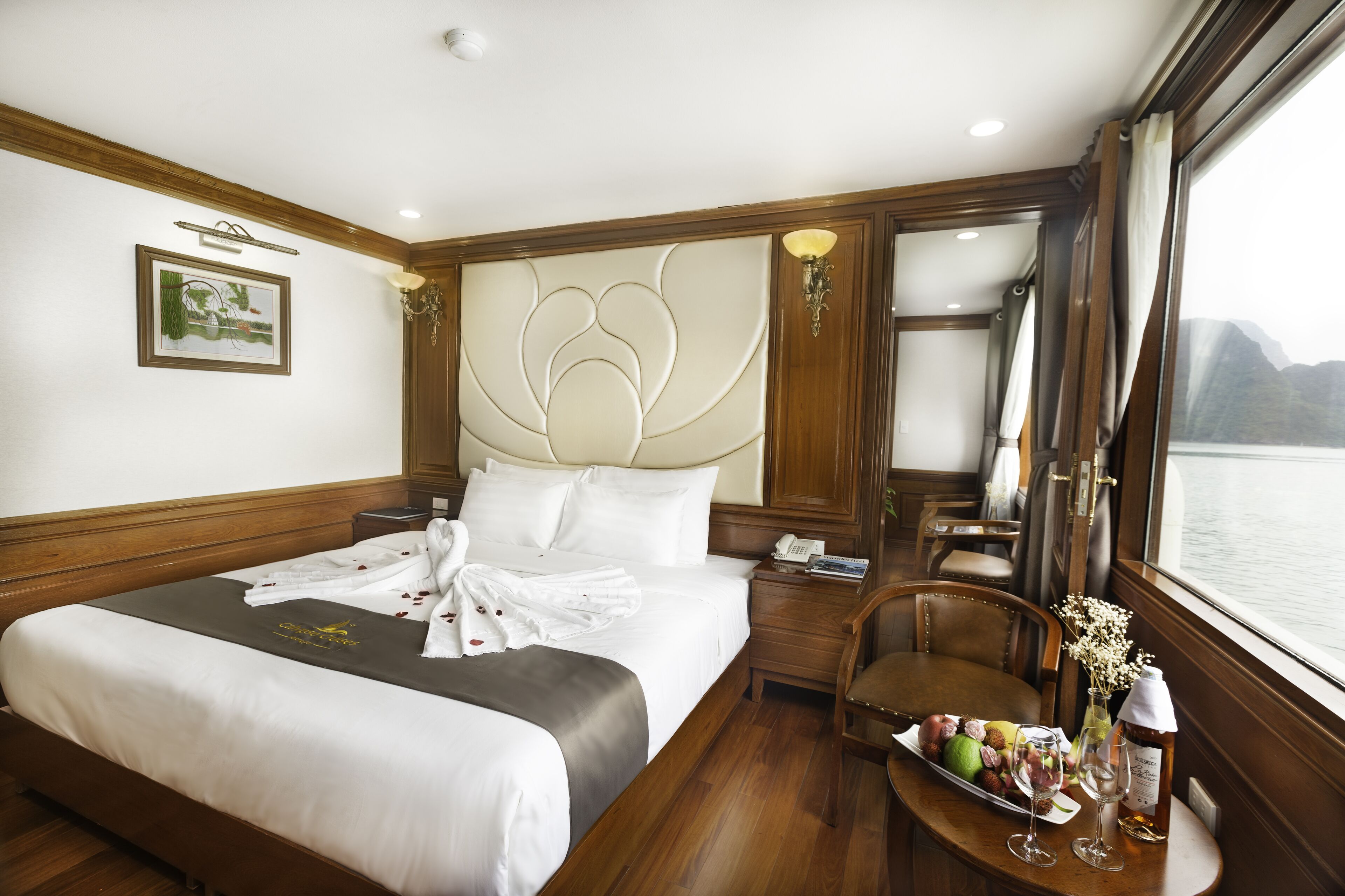 Une chambre d’hôtel avec un grand lit, une table de chevet avec une lampe, une chaise, une petite table avec un plateau de fruits et des verres, et une fenêtre donnant sur l’eau.