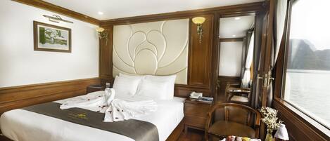 Une chambre d’hôtel avec un grand lit, une table de chevet avec une lampe, une chaise, une petite table avec un plateau de fruits et des verres, et une fenêtre donnant sur l’eau.