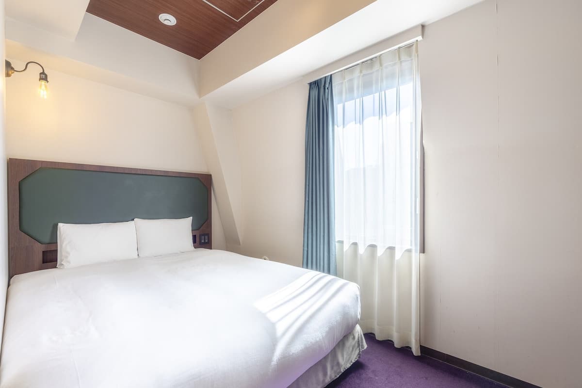 A hotel room with a large bed, a wooden headboard, a window with sheer curtains, and a purple carpet.