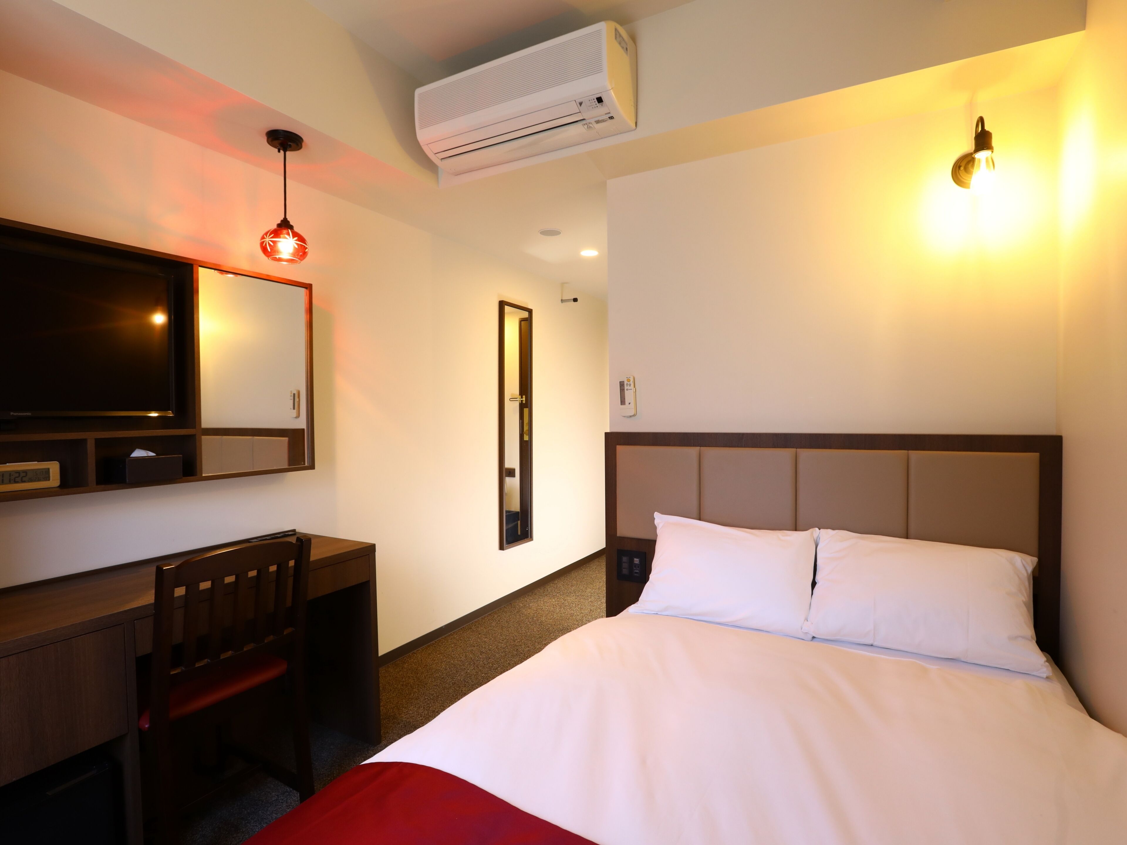 A hotel room with a bed, a desk, a chair, a TV, an air conditioner, and a wall-mounted light.