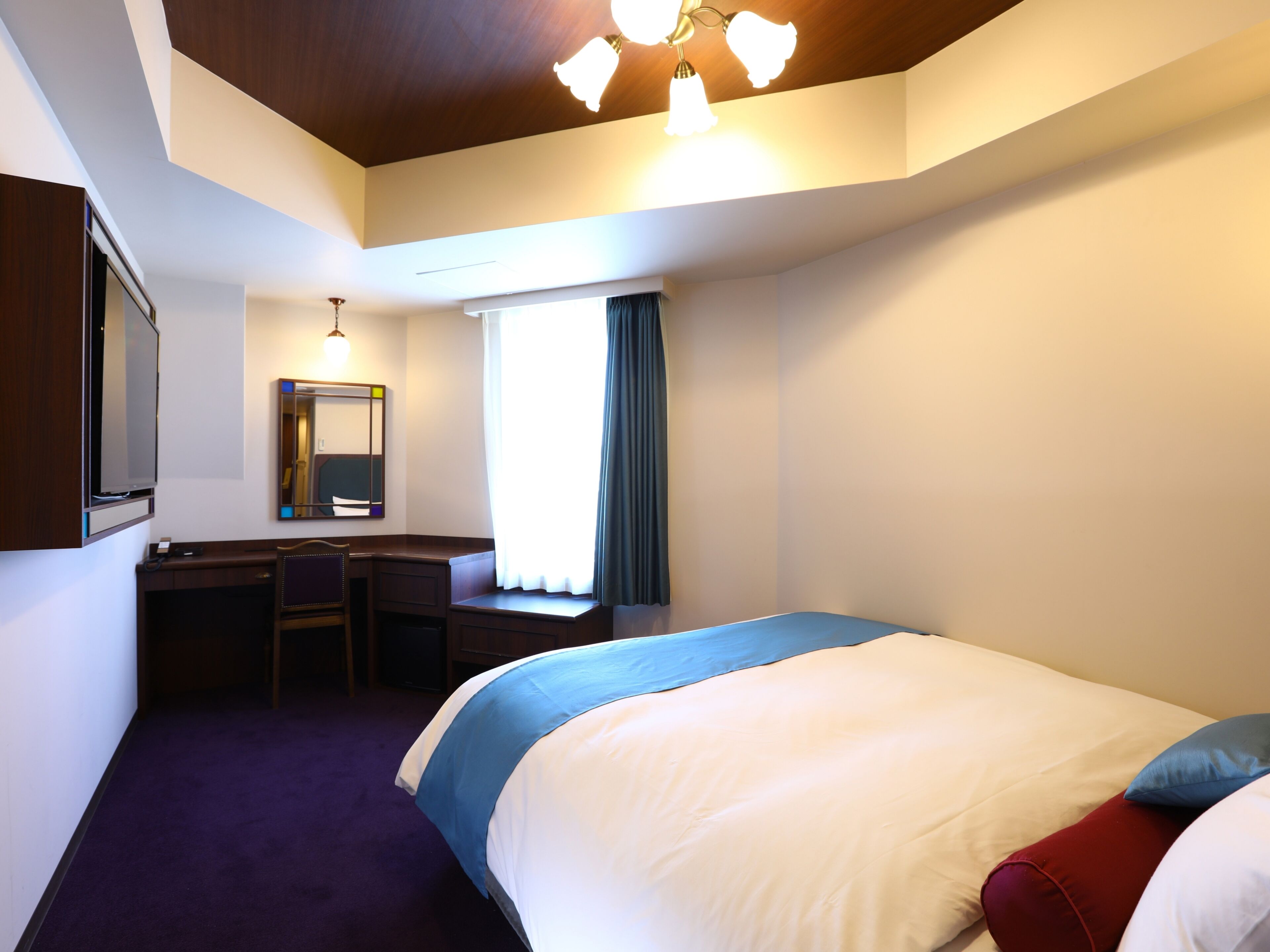 Una habitación de hotel con cama, escritorio, silla, televisión, espejo y ventana con cortinas.
