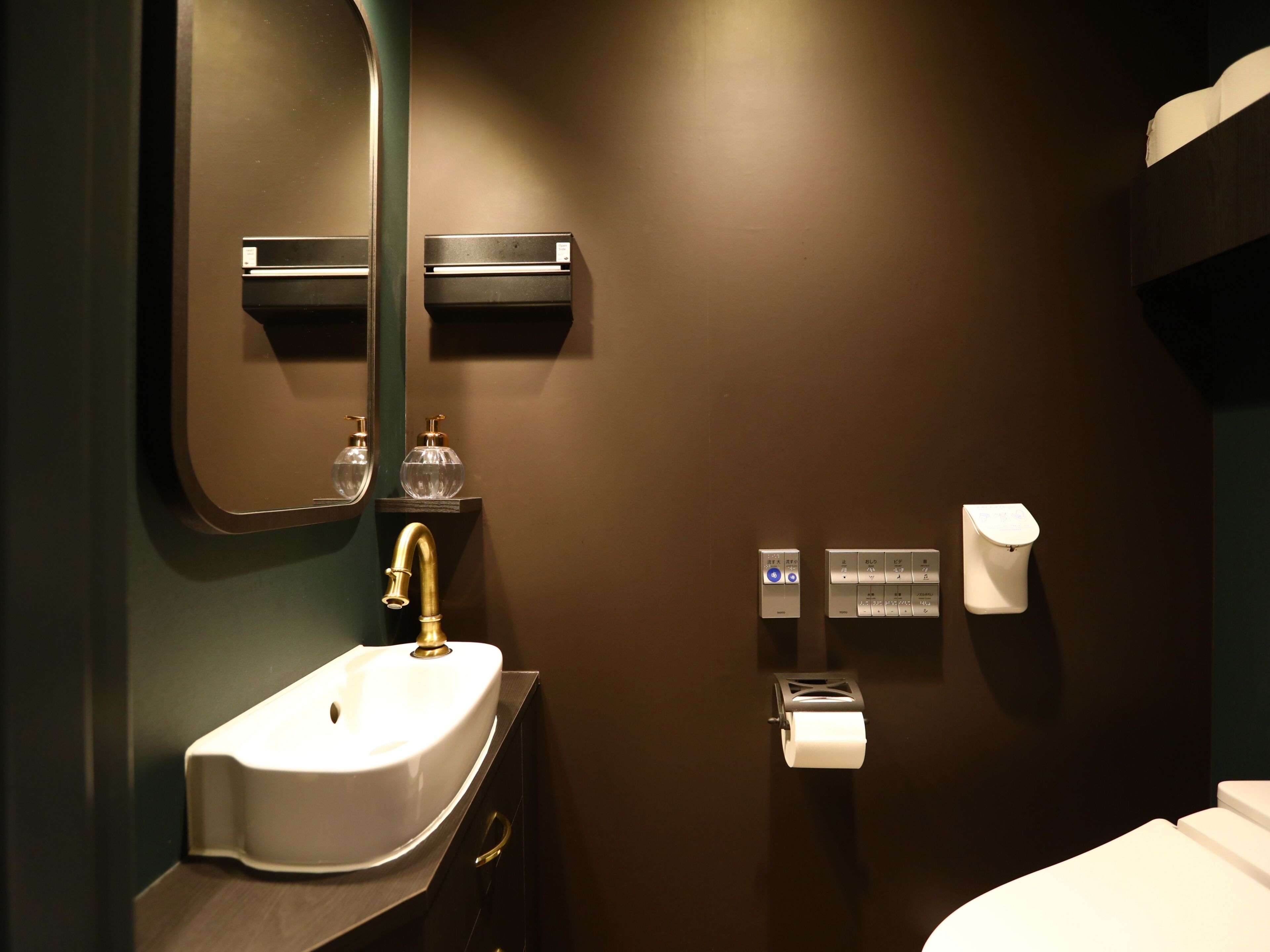 Un bagno moderno con un lavandino bianco, un rubinetto dorato e uno specchio sopra. Si vedono due dispenser di sapone su uno scaffale, un portasciugamani e un water.
