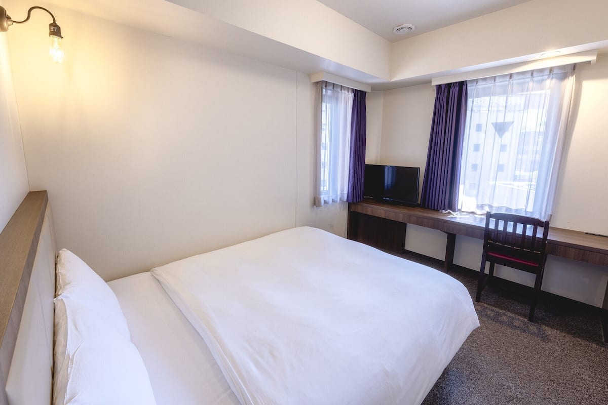 A hotel room with a bed, a desk with a television, and a window with curtains.