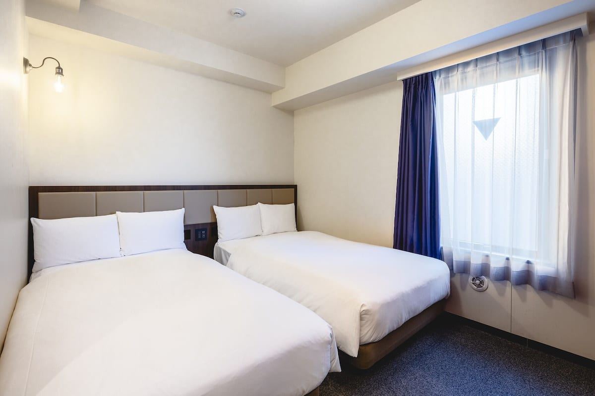 A hotel room with two beds, a ceiling light, a window with curtains, and a carpeted floor.