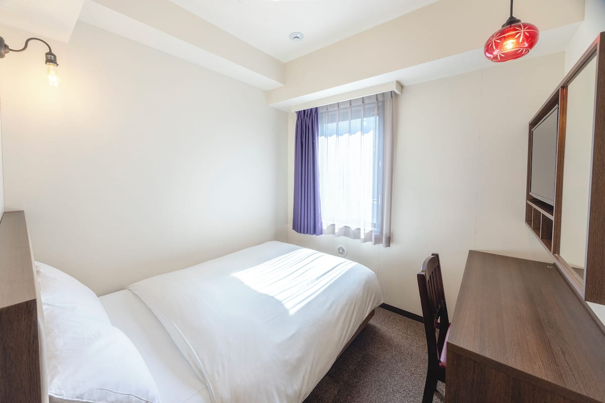 A hotel room with a single bed, a wooden desk, a chair, a window with purple curtains, and a red pendant light.