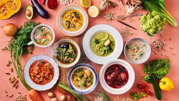 Variedad de tazones de sopa con diferentes ingredientes, rodeados de verduras frescas y hierbas sobre un mantel rosa.