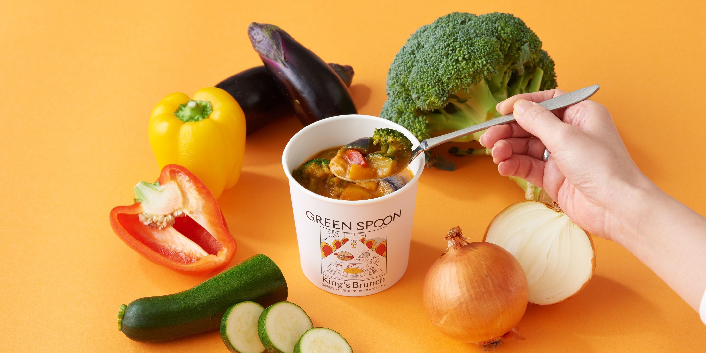 A hand holding a spoon over a bowl labeled "GREEN SPOON King's Brunch" with vegetables and a bell pepper on an orange background.