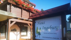 Letrero de 'Haus Ingeburg' con una dirección web, un logotipo con árboles y un edificio de madera con balcón y macetas con flores.
