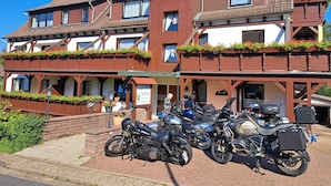 Un edificio tradicional europeo con fachada de varios pisos, balcones de madera con macetas de flores y una fila de motocicletas estacionadas en la calle.