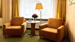 Una habitación de hotel con dos sillones, una mesita con un florero, una lámpara de pie y una ventana con cortinas.