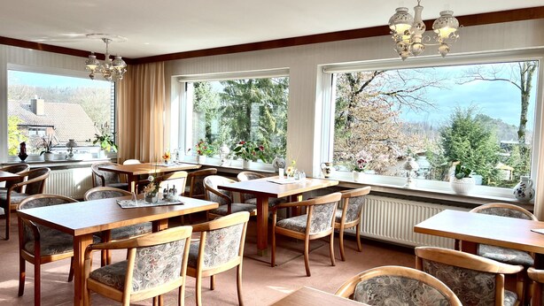 Un comedor con mesas y sillas de madera, amplias ventanas con vistas a árboles y edificios, e iluminación decorativa.