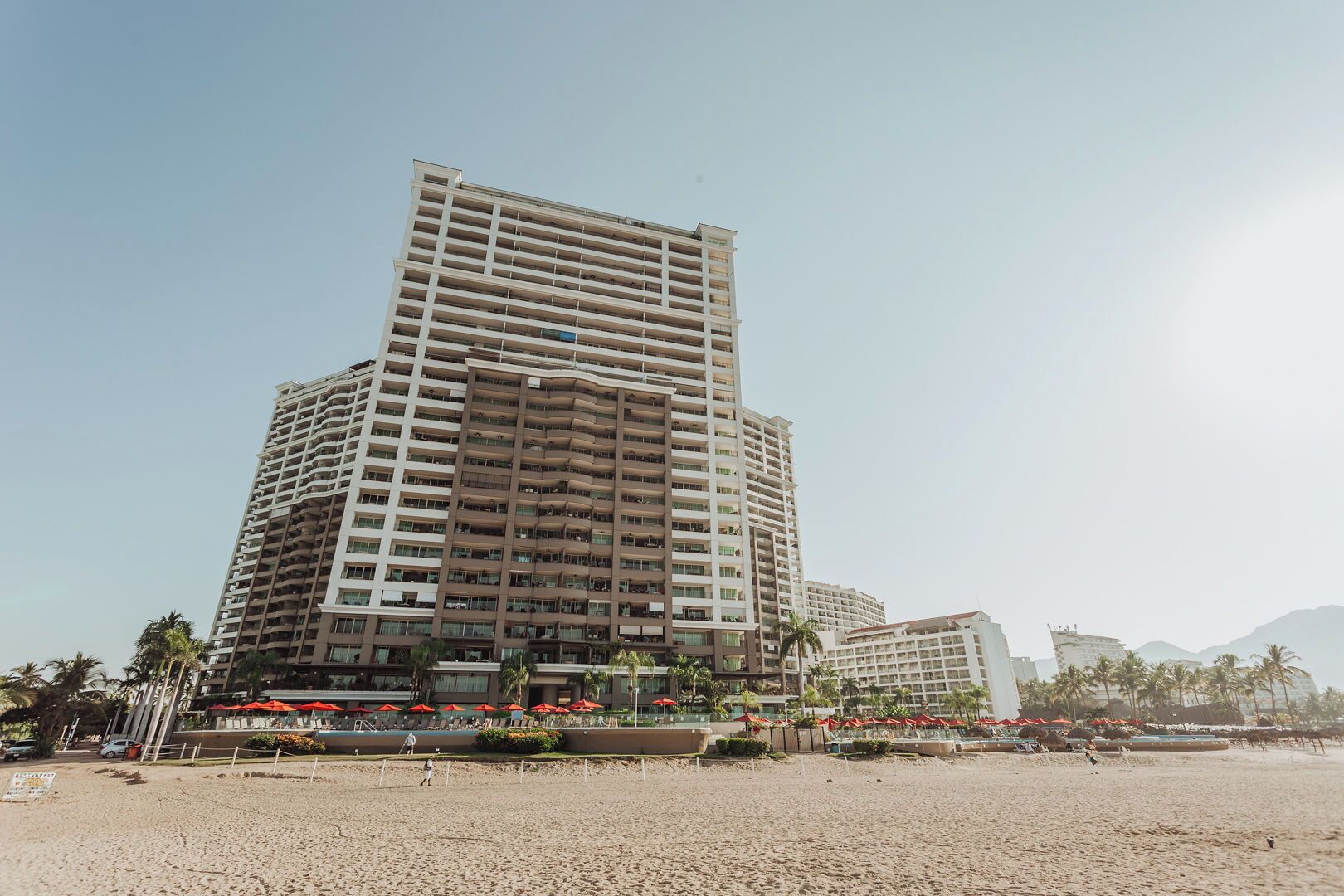 Un hôtel à plusieurs étages au design moderne, doté de balcons à chaque niveau, situé près d’une plage bordée de palmiers et parsemée de parasols.