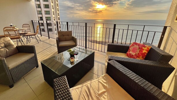 A balcony with wicker furniture, a glass table, and a view of the sunset over the sea. - ☼Hypnotizing sea view on 17th floor ~Prime location in town ~Family getaway (Puerto Vallarta)