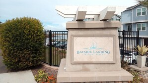 Skilt for Bayside Landing Imperial Beach med frit udsyn over området, herunder et hegn, parkerede biler og et lille træ.