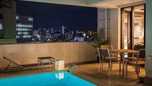 Een zwembad op het dak met uitzicht over de stad in de nacht, inclusief een tafel met stoelen en een ligstoel bij het zwembad.