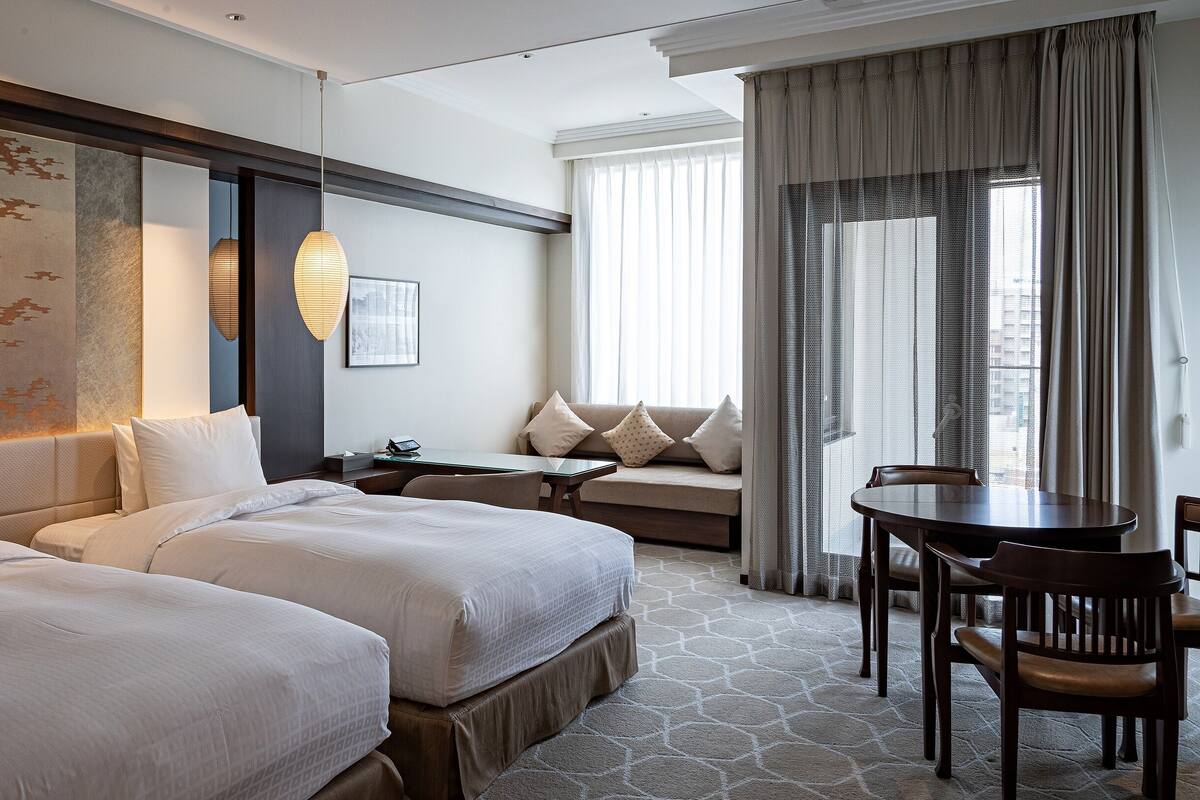 Een hotelkamer met twee bedden, een bank, een eettafel en een groot raam met gordijnen.
