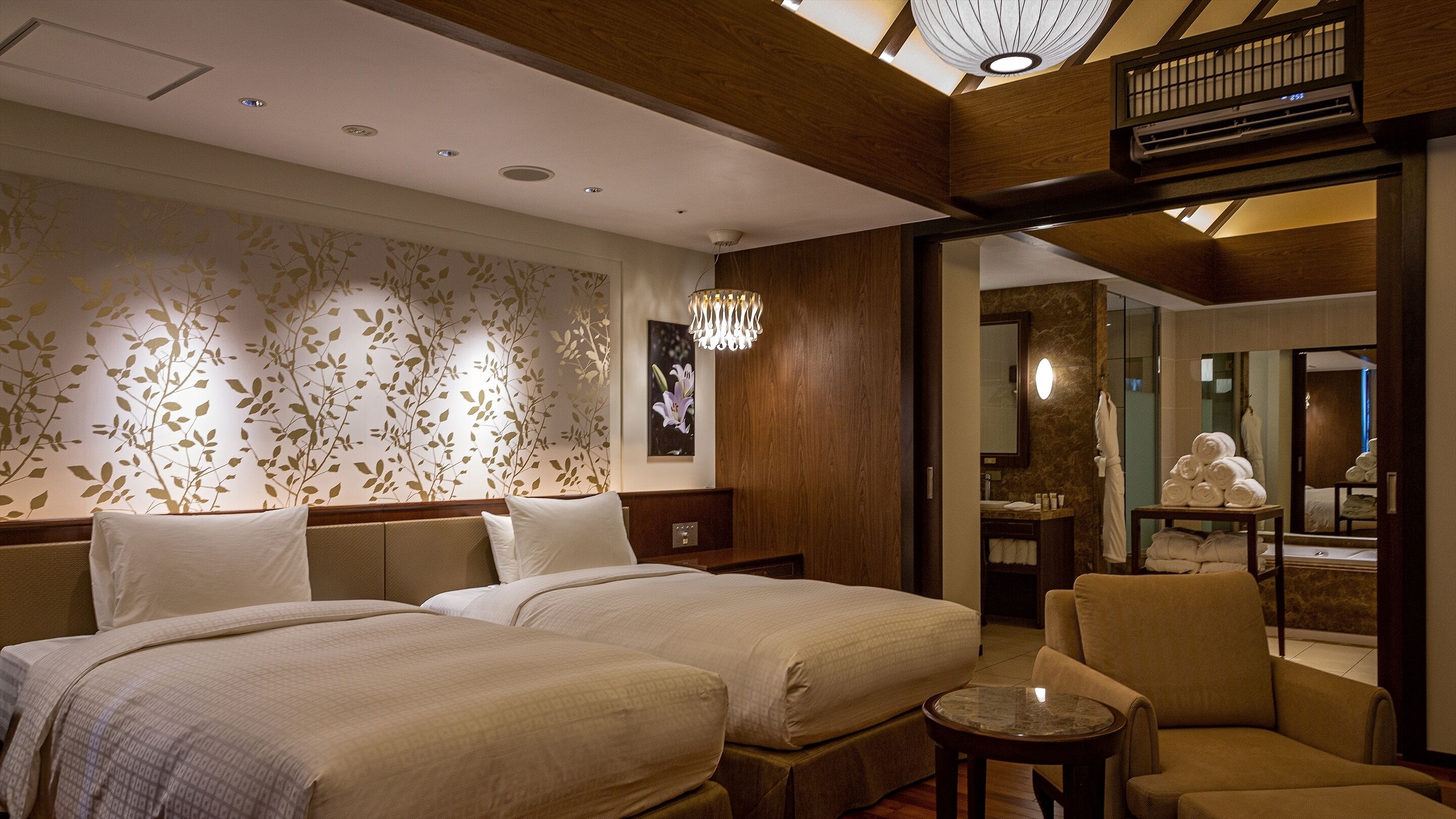 Une chambre d’hôtel avec deux lits, un coin salon comprenant un fauteuil, une petite table, un miroir et une salle de bain visible à travers une porte ouverte.