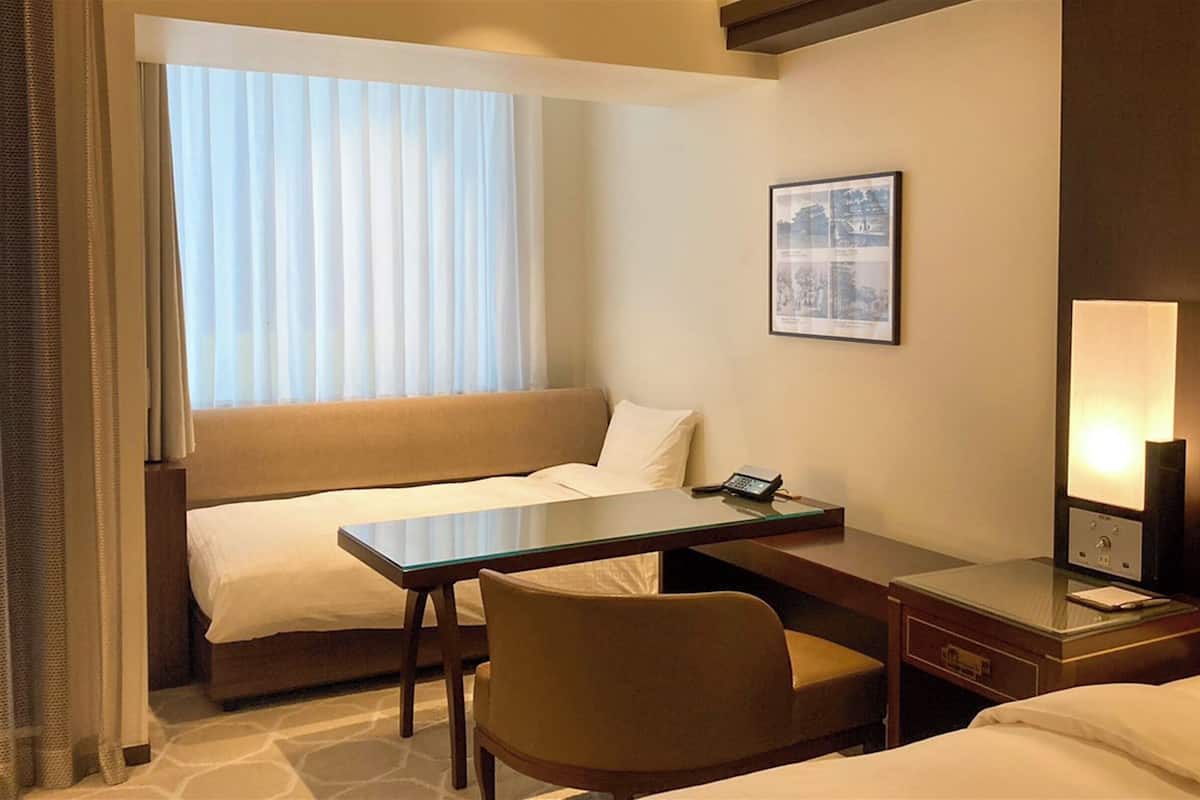 Een hotelkamer met twee bedden, een bureau, een stoel, een nachtkastje, een lamp en een ingelijst schilderij aan de muur.
