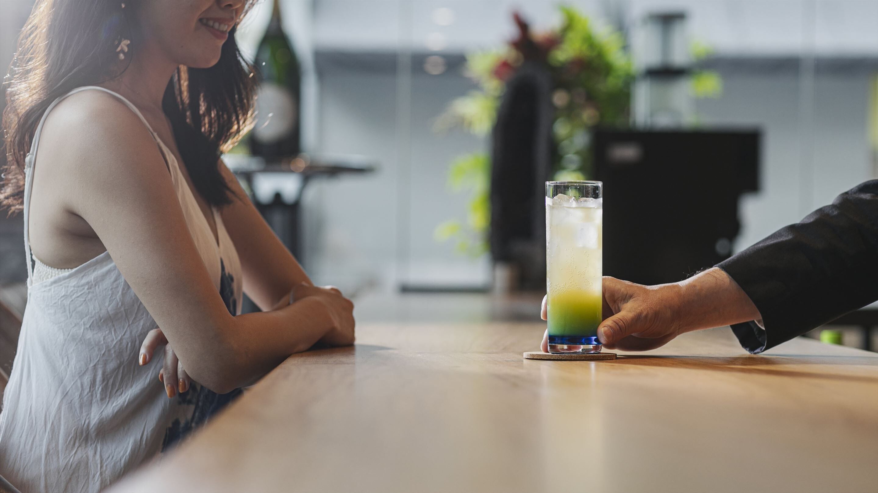 A person holding a tall glass with a layered drink, standing at a bar with a wooden surface.