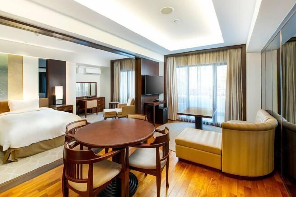 Een hotelkamer met een bed, een eettafel, een sofa, een televisie en een groot raam met gordijnen.