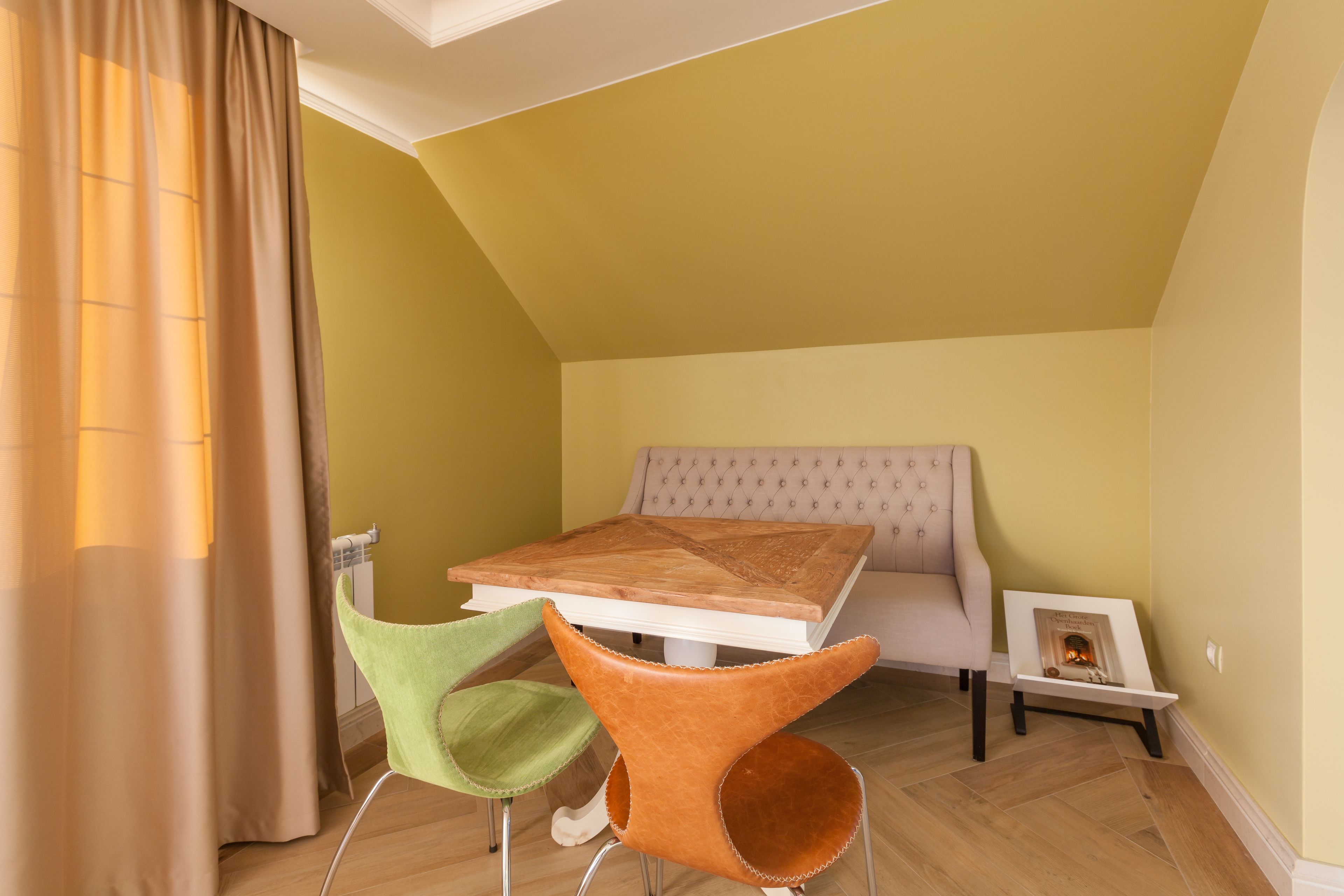 Une pièce avec un plafond en pente, une table en bois, une chaise verte et une chaise orange, un canapé et une petite cheminée.