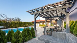 A covered outdoor seating area with wicker furniture, a pool, and a stone wall in the background. - Villa Ciconia (Veliko Tarnovo)