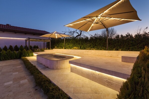 Un espace extérieur bien éclairé, avec un patio, un foyer intégré, des sièges et de grands parasols pour créer de l’ombre.