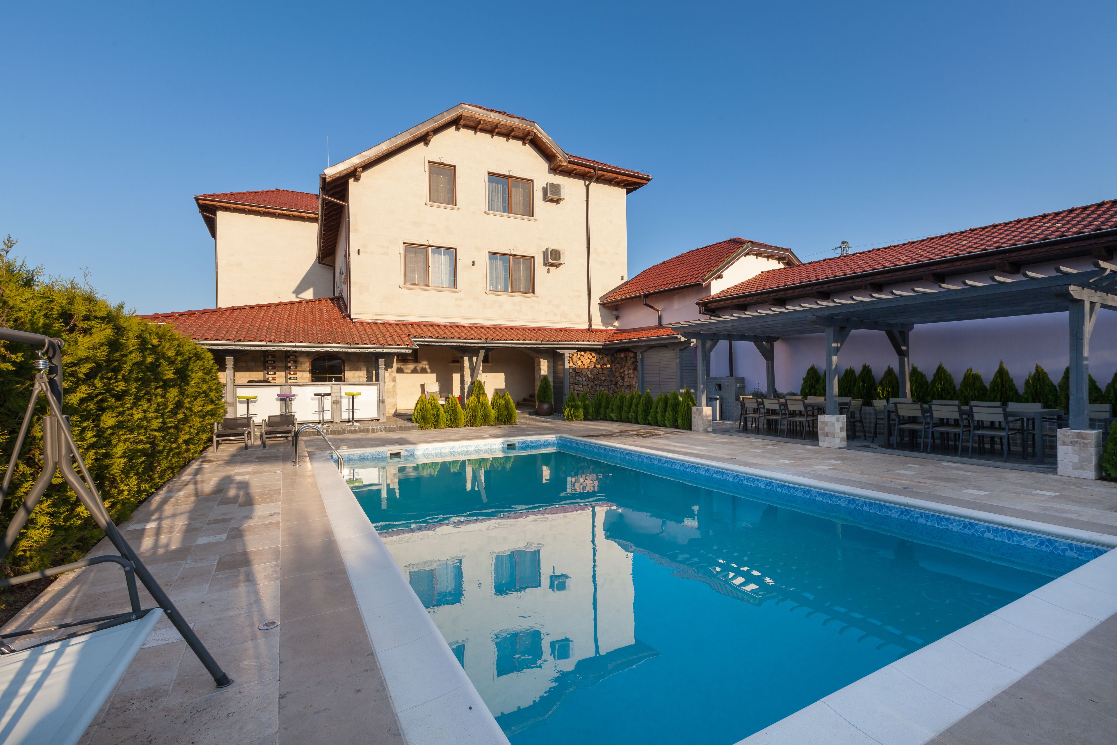 A swimming pool with a tiled deck, a covered patio area, and a building with a red-tiled roof.