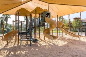 A playground with multiple slides and climbing structures, surrounded by a sandy area and shaded by a large canopy.