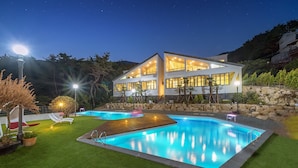 A modern two-story house with a swimming pool, surrounded by a well-maintained lawn and a rocky hillside. - Ruscello Pension (Geoje)