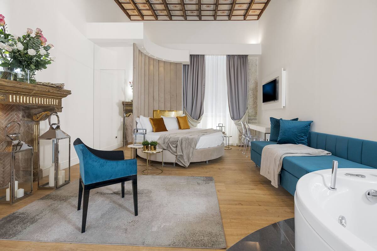 A modern hotel room with a bed, a blue sofa, a white bathtub, and a seating area with a chair and a small table.