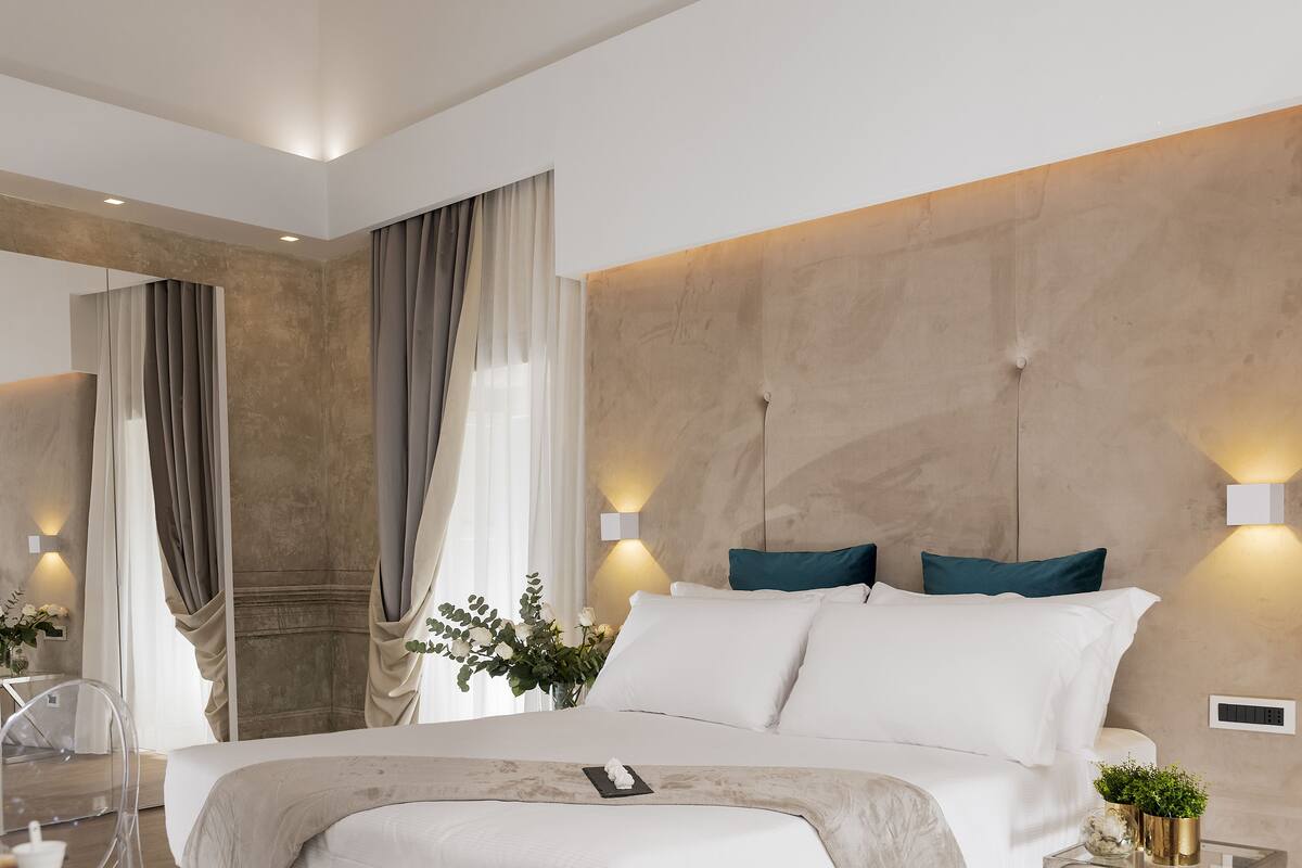 A modern hotel room with a large bed, a bedside table with a vase of flowers, and a wall-mounted light fixture.