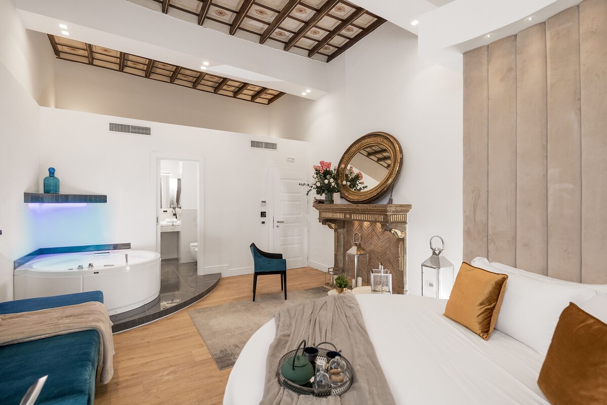 A modern hotel room with a large bed, a sitting area with a blue chair, a bathroom with a bathtub, and a fireplace.