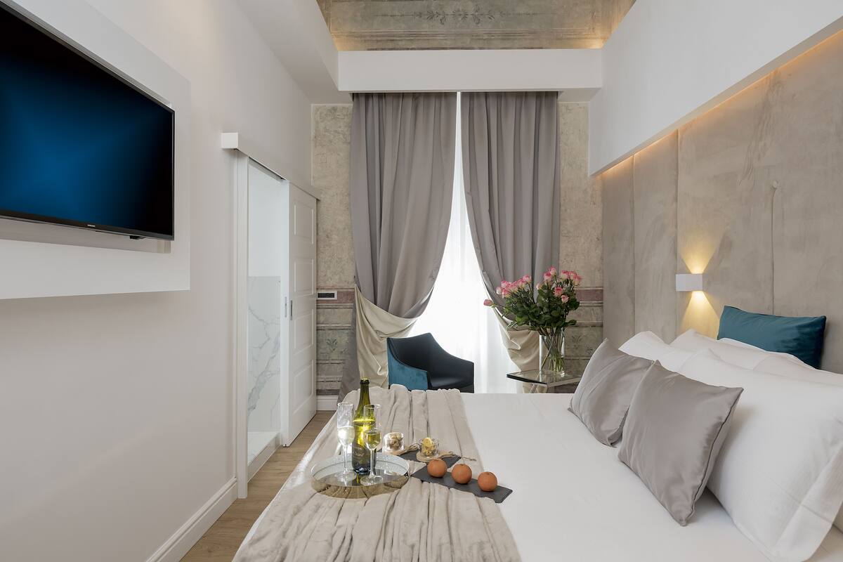 A modern hotel room with a large bed, a flat-screen TV mounted on the wall, a dining table with a bottle of wine, and a vase with flowers.