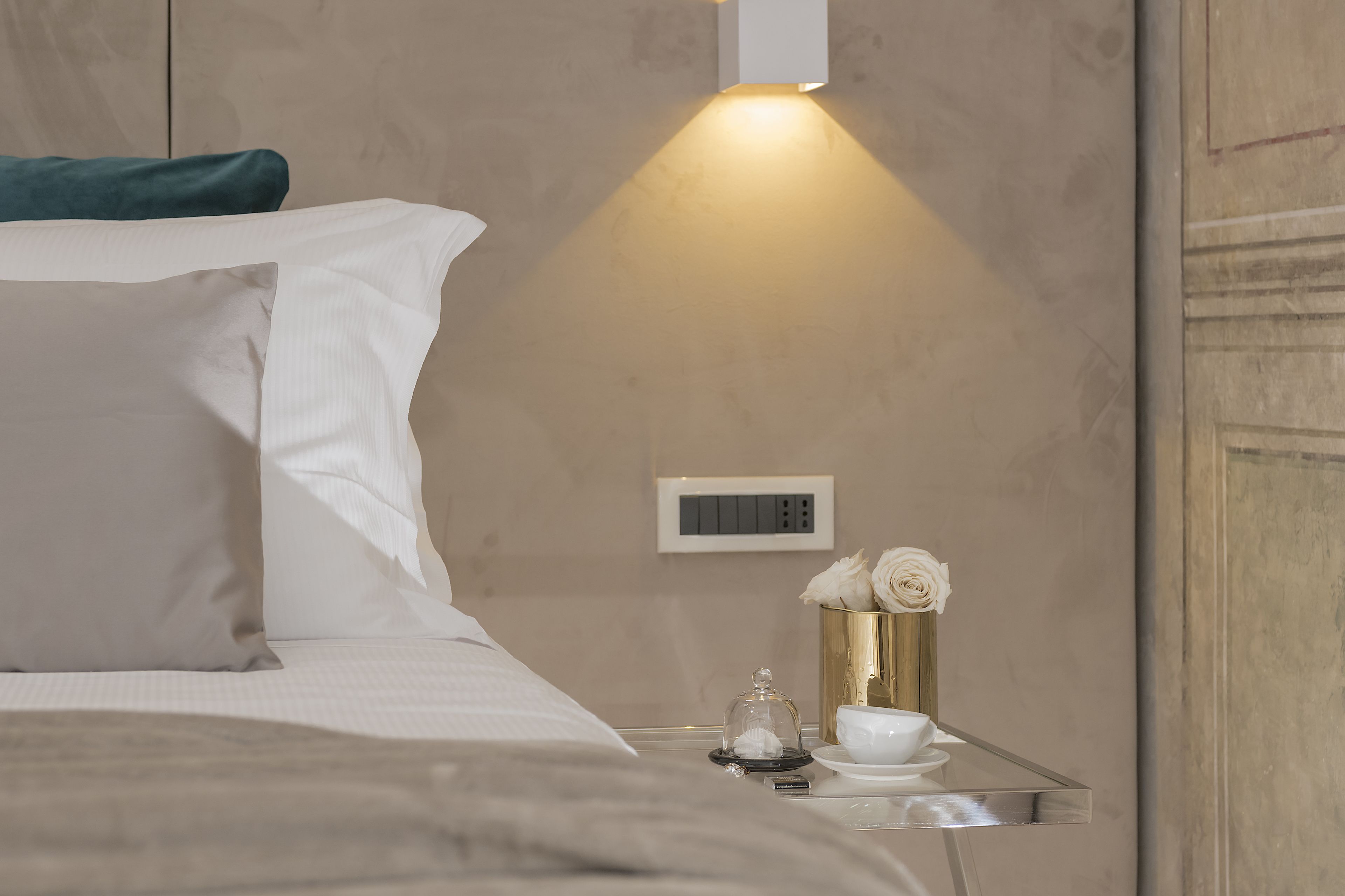 A neatly made bed with white linens, a bedside table with a glass top, a small vase with white flowers, and a wall-mounted thermostat.