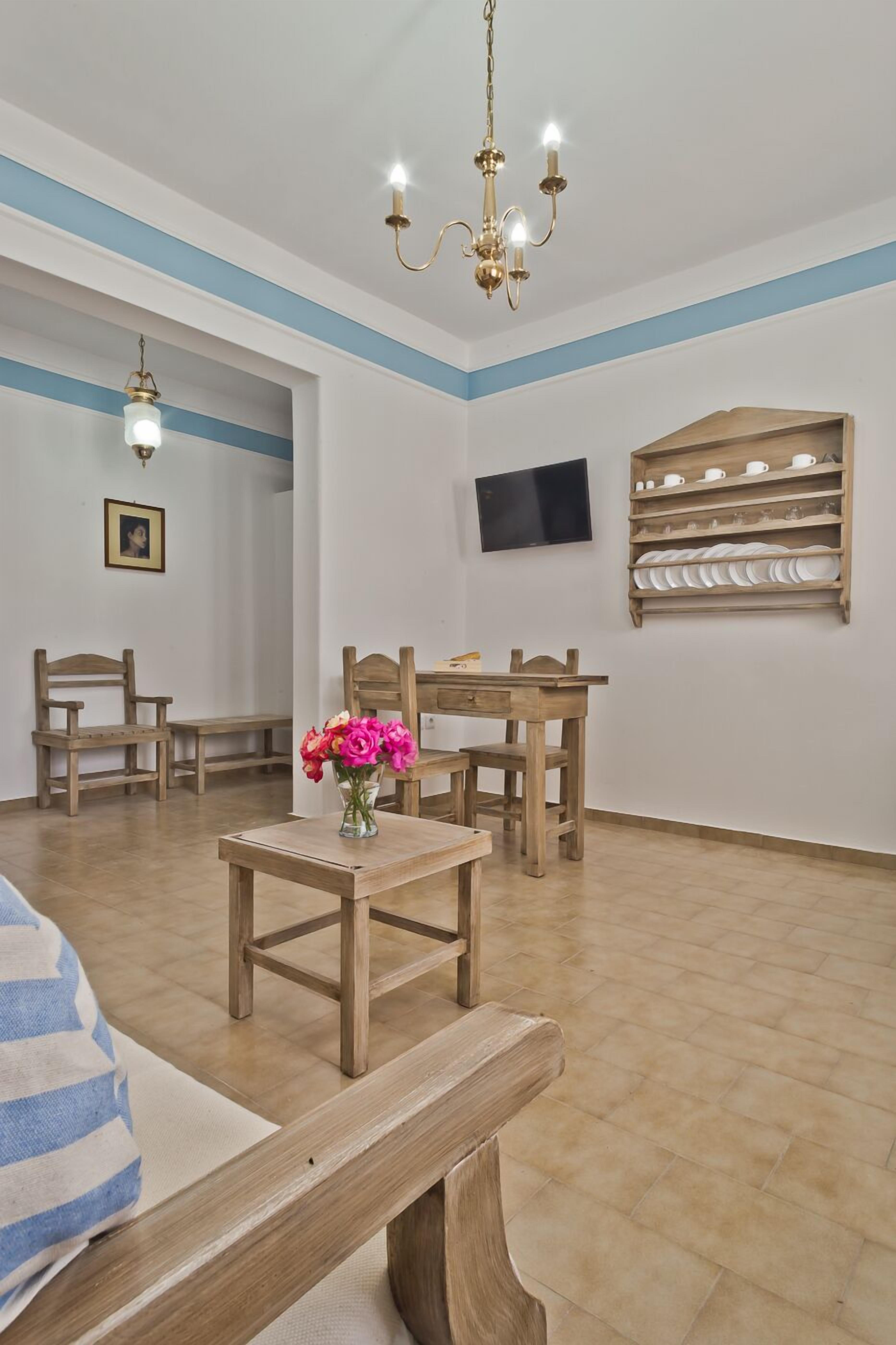 A room with a wooden dining table, chairs, a vase with flowers, a television on the wall, and a shelf with plates.