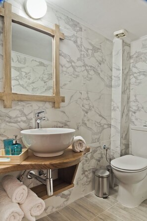 A bathroom with a marble-tiled wall, a wooden vanity with a sink, a mirror, and a toilet. - Philoxenia (Karpathos)