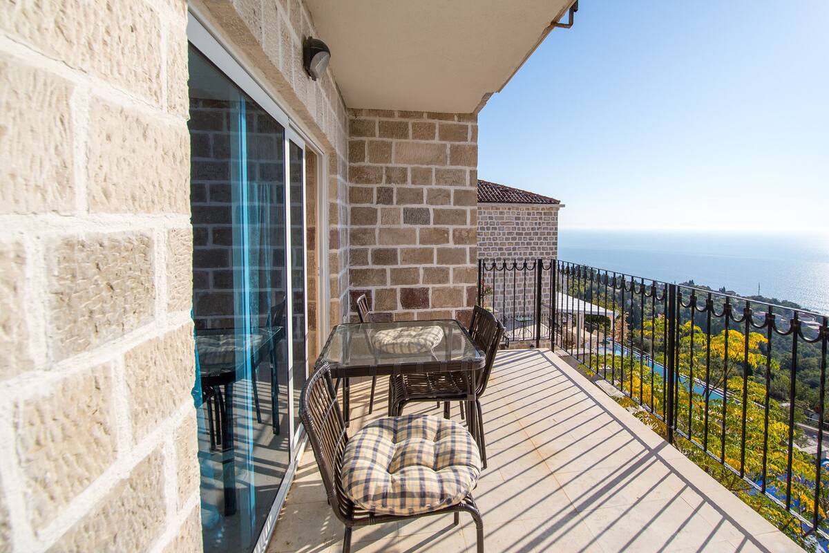 A balcony with a glass table and chairs, a stone wall, and a view of the sea.