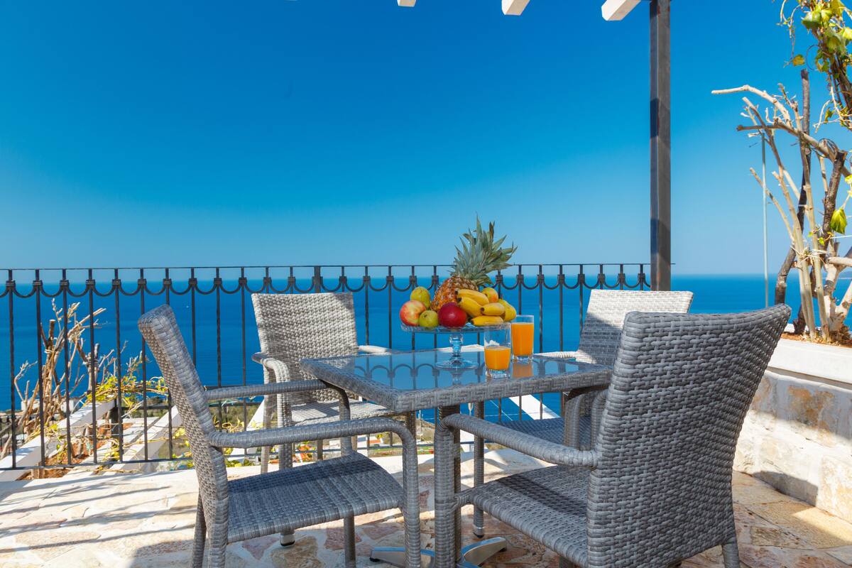 A wicker table and chairs set on a balcony with a fruit bowl and drinks, overlooking a clear blue sea.