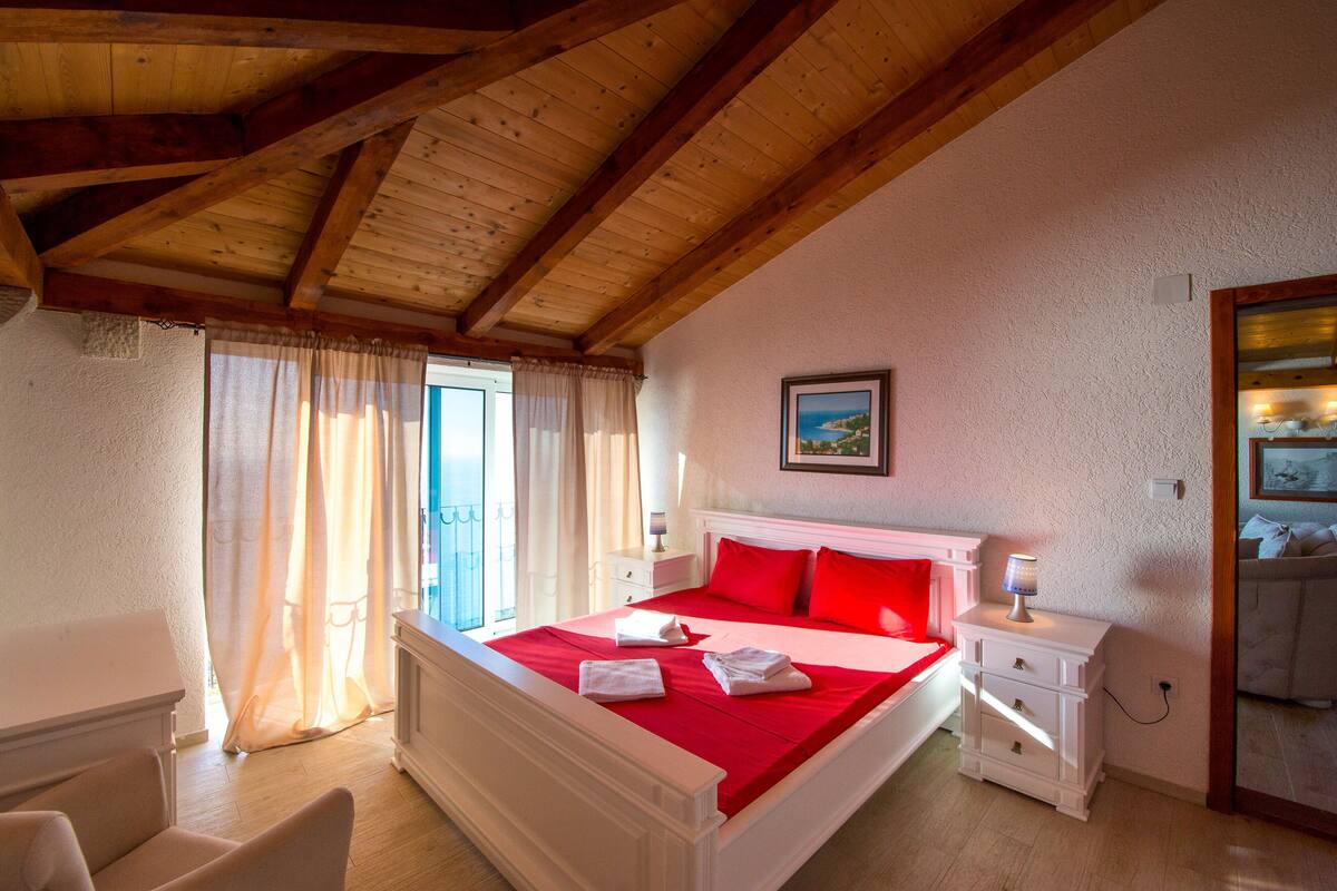 A bedroom with a wooden ceiling, a bed with red bedding, a nightstand, a lamp, and a painting on the wall.