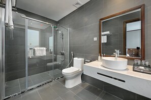 Un baño moderno con ducha de vidrio, inodoro blanco, lavamanos grande con espejo y paredes de azulejos oscuros.