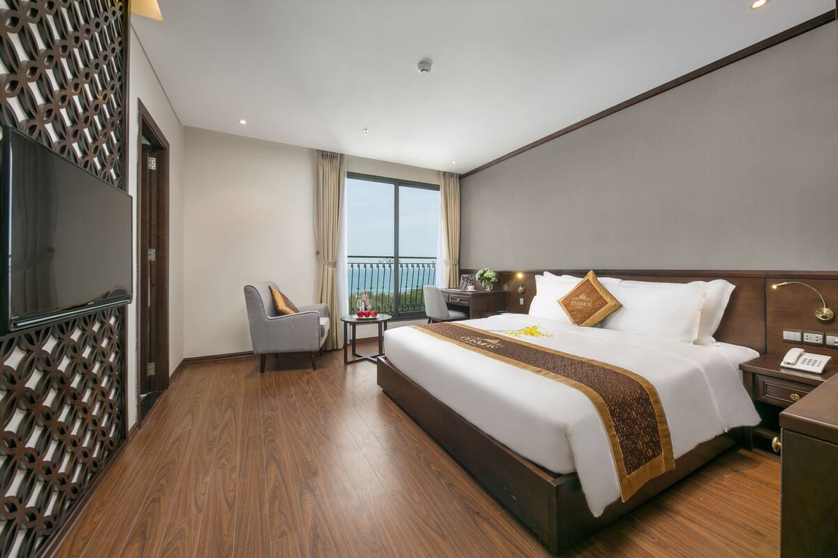 A hotel room with a large bed, a desk, a chair, a TV, and a balcony with a view.
